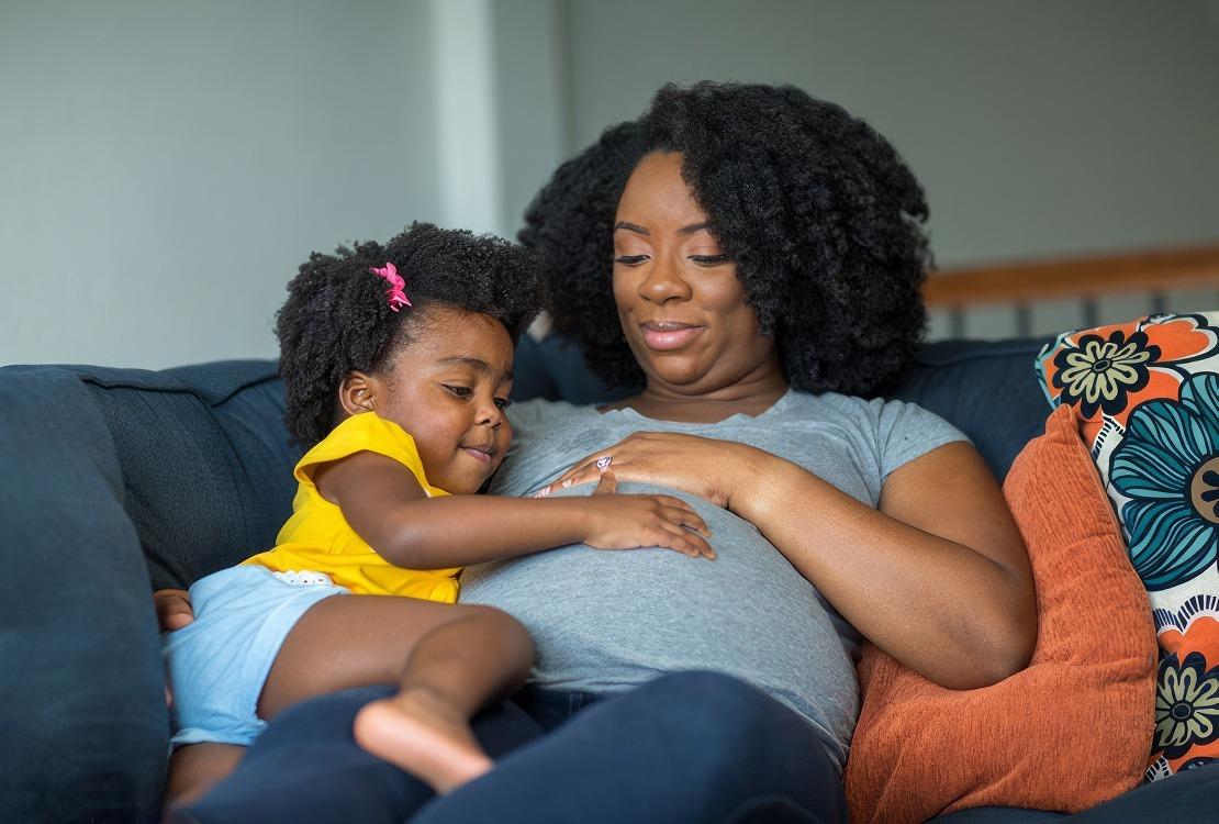 A pregnant mom sitting on the couch with her daughter feeling her belly